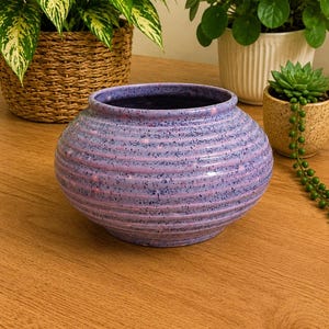 May include: A round, speckled purple ceramic planter with horizontal ridges. The interior is dark purple. The planter sits on a wooden surface, with various potted plants in the background, including a woven basket planter.