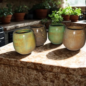 May include: Four small, rounded ceramic vases in shades of green, brown, and tan sit on a countertop. The vases have a textured, mottled appearance and a dark rim. The countertop is a speckled brown and tan.