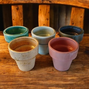 May include: Six small ceramic cups in various colors, including blue, green, and pink, with speckled patterns. The cups are arranged on a wooden surface.