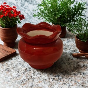 May include: A glossy, red ceramic pot with a ruffled rim, ideal for plants. The pot sits on a speckled surface, with other small terracotta pots holding greenery and red flowers in the background. A wooden cutting board is also visible.