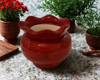 May include: A glossy, red ceramic pot with a ruffled rim, ideal for plants. The pot sits on a speckled surface, with other small terracotta pots holding greenery and red flowers in the background. A wooden cutting board is also visible.