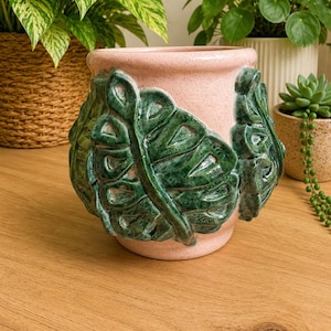 May include: A pink ceramic planter with a speckled finish and a green, raised leaf design. The planter is cylindrical and sits on a wooden surface. The design features large, detailed leaves, adding a touch of nature to the decor.