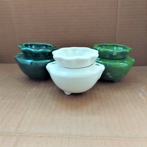 May include: A collection of small ceramic pots in shades of green and white. The pots have a rounded base and a scalloped rim. The white pot has three small feet. The green pots have a speckled finish. These decorative pots are suitable for small plants.