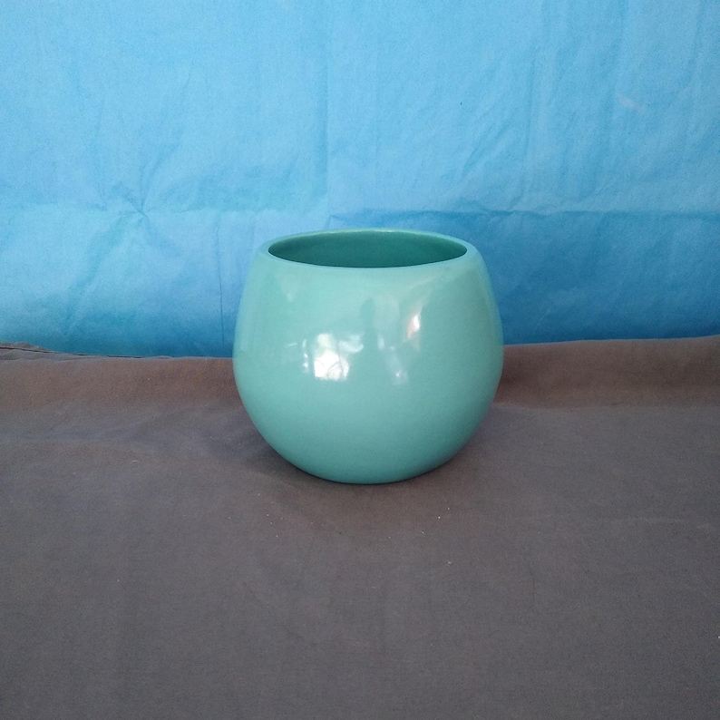 May include: A light blue ceramic planter with a rounded shape. The planter is smooth and glossy.