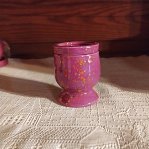 May include: A small, purple ceramic container with a lid. The container has a speckled design with orange and yellow dots. It has a rounded body and a pedestal base. The container is sitting on a white, patterned tablecloth.