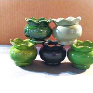 May include: A collection of six small, ceramic pots in various shades of green, including a deep forest green, a lighter lime green, and a pale green. Each pot has a ruffled, scalloped rim and a rounded body. The pots are stacked and arranged on a light surface.