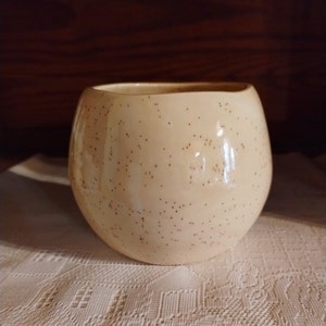 May include: A cream-coloured ceramic mug with a speckled brown glaze. The mug has a rounded shape and a wide opening.