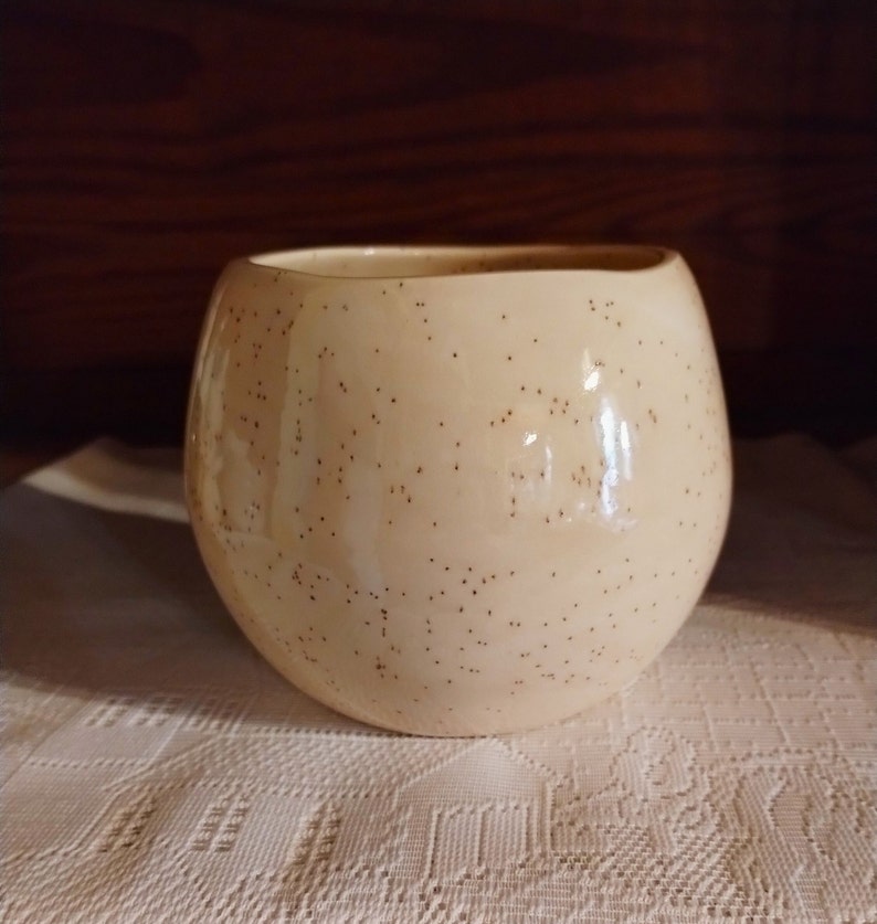 May include: A cream-coloured ceramic mug with a speckled brown glaze. The mug has a rounded shape and a wide opening.