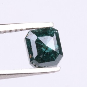 May include: A dark teal gemstone with an emerald cut is held by silver tweezers. The gem has a square shape with beveled edges, reflecting light and showcasing its facets. The background is plain white.