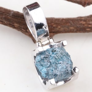 May include: A silver pendant with a rough, light blue gemstone. The pendant is set in a silver frame and hangs from a silver bail. The background is white with a brown twig.