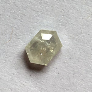 May include: A small, light yellow-toned diamond with a unique hexagonal shape. The gemstone has a faceted surface that reflects light, creating a sparkling effect. The diamond is set against a plain, off-white background.