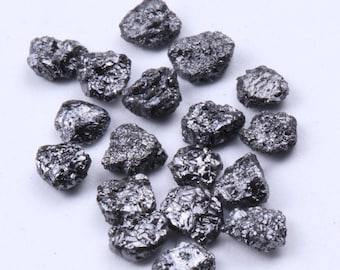 May include: A collection of rough, uncut black diamonds. These raw gemstones have a textured, irregular surface and a dark, metallic sheen. The diamonds are scattered on a white surface.