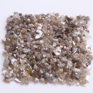 May include: A collection of small, irregularly shaped, raw gemstones. The stones are primarily shades of brown, tan, and clear, with some having a reflective, iridescent quality. The gemstones are arranged in a rectangular shape against a white background.