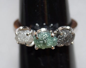 May include: A silver ring with three rough diamonds. The diamonds are white, green, and black.