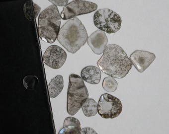 May include: A collection of irregularly shaped, translucent gemstones with internal patterns of gray and white. The stones are scattered on a white surface, with a black, rectangular object in the corner.