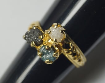 May include: A gold ring featuring a cluster of rough-cut gemstones in gray, white, and teal. The ring has an ornate, filigree design on the band, adding a touch of elegance to the unique gemstone arrangement.