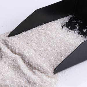 May include: A black scoop containing small, clear, glitter-like particles. The particles are spilling onto a white surface. The scoop is angled, and the particles are scattered.