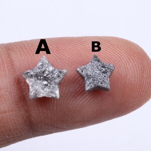 Pode incluir: Dois pequenos objetos cinzentos em forma de estrela, rotulados A e B. As estrelas têm uma superfície áspera e texturizada e são seguradas entre os dedos. As estrelas são provavelmente pedras preciosas ou elementos decorativos.