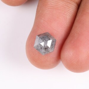 May include: A hexagonal, grey diamond with facets, held between two fingers. The gemstone has a speckled appearance and reflects light, showcasing its cut and clarity. The diamond is a unique shape, perfect for jewellery.