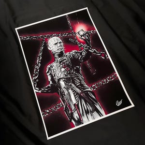 May include: A black and white illustration of a character from the horror film Hellraiser, surrounded by chains. The artwork features the character with pins in their head, holding a puzzle box, with a red background.