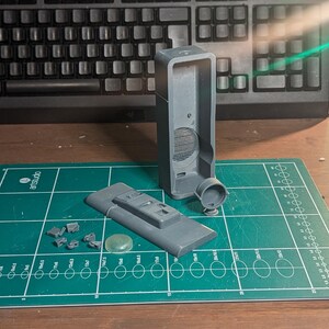 May include: A disassembled gray electronic device with various components, including a rectangular body, a circular piece, and a rectangular base with buttons. The parts are arranged on a green cutting mat with a keyboard in the background.