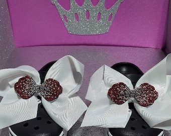 May include: A pair of black clogs with white trim, each decorated with a large white bow accented with a red rhinestone bow.