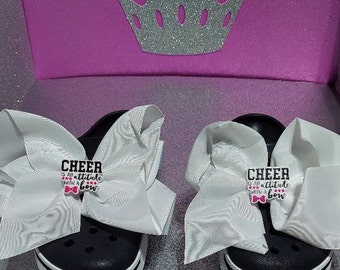 May include: A pair of black and white slip-on shoes with white bows and a pink and black charm that says "Cheer is an attitude with a bow".
