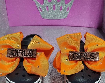 May include: A pair of black clogs with orange bows and rhinestones. The bows have a brown, glittery "GIRLS" embellishment.
