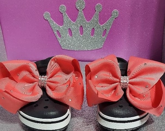 May include: A pair of black clogs with a white stripe around the sole. Each clog has a large coral-colored bow with a rhinestone center.