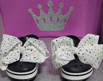 May include: A pair of black clogs with white trim and a large white bow with colorful rhinestones on each shoe.