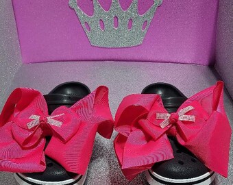 May include: A pair of black clogs with white trim and large pink bows with rhinestones. The bows are tied around the clogs and are a statement piece.