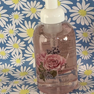 May include: A clear plastic bottle of foaming hand soap with a white pump dispenser. The bottle has a floral design with a pink rose and purple flowers. The label reads "Pure Passion™ Soaps and Lotions Rose & Lilac Foaming Hand Soap 500 ml. 16.9 fl. oz."