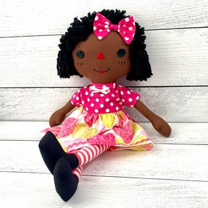 Raggedy Ann Doll, with dark brown skin tone and black hair, wearing pink dot and heart dress