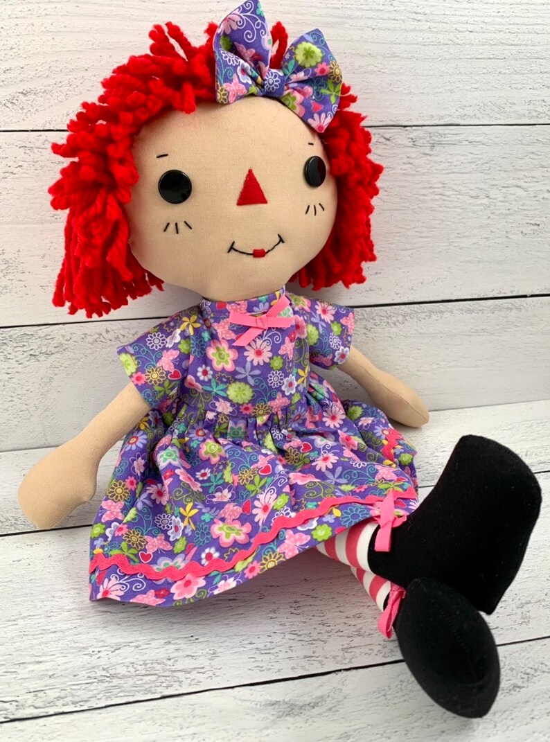 Raggedy Ann style doll is dressed in a removable floral light purple dress with matching hair bow. 
She has light tan skin tone with red hair. Recommended for ages 3 and up.
Doll Sizes:
Small 13 inches
Medium 16 inches
Large 20 inches