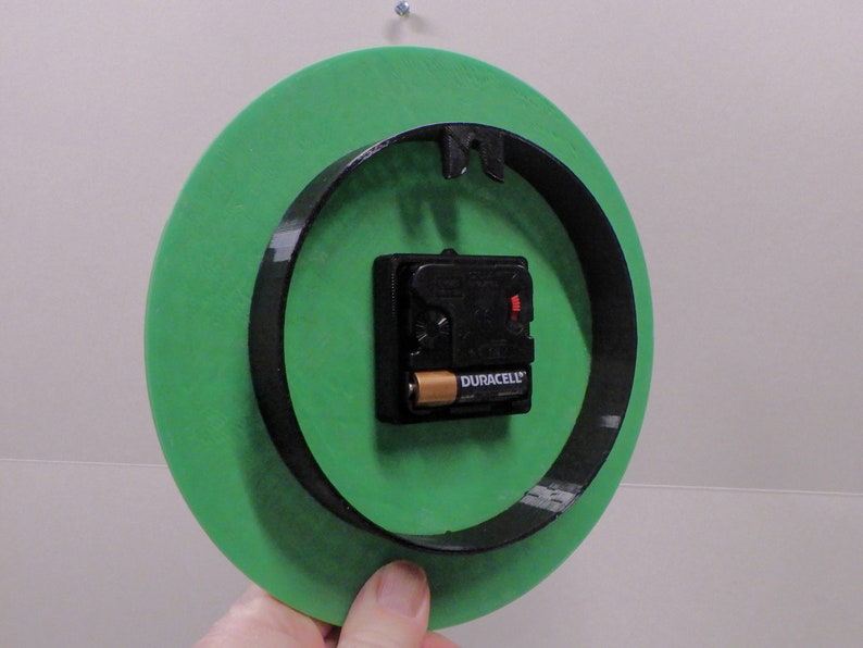 以下が含まれることがあります： 黒い縁取りのある丸い緑色の時計の裏側。Duracellのバッテリーが付いた黒い時計機構が取り付けられています。時計は壁に掛けられています。