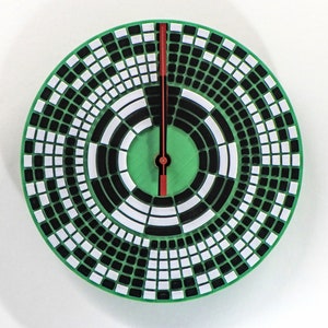 以下が含まれることがあります： ユニークなデザインの丸い緑色の時計。文字盤は、同心円状に配置された白と黒の正方形のパターンです。赤い針が時間を指しています。時計の直径は約25cmです。