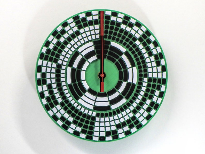 以下が含まれることがあります： ユニークなデザインの丸い緑色の時計。文字盤は、同心円状に配置された白と黒の正方形のパターンです。赤い針が時間を指しています。時計の直径は約25cmです。
