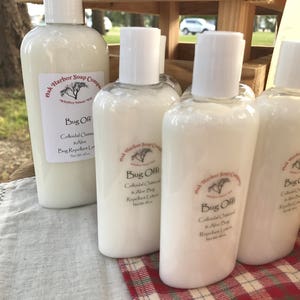 May include: Several white plastic bottles of "Bug Off" lotion are arranged on a red and white checkered cloth. The bottles have white caps and labels with text. The lotion is from Oak Harbor Soap Company.