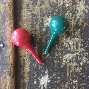 May include: Two wooden ornaments, one red and one green, with rounded tops and long, thin stems. The ornaments are on a textured, brown surface.