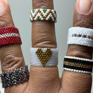 May include: A hand model wearing six different beaded rings. The rings are made with various colors and patterns, including red, white, green, gold, and black.