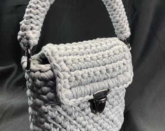 May include: A light gray crocheted handbag with a rounded handle and a rectangular body. The bag features a flap closure with a silver-colored clasp. The crochet work is visible throughout the bag, creating a textured appearance. The bag is set against a dark background.