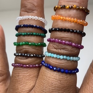 May include: A set of ten beaded rings in various colors, including clear, orange, black, green, red, blue, and turquoise. The rings are stacked on a person's finger.