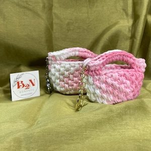 May include: Two small, handmade crochet bags in pink and white, with matching handles and gold-colored chain accents. A small square card with the text "B₂N GONZALO" is to the left of the bags. The bags are on a gold-colored fabric.