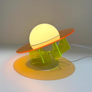 May include: A table lamp featuring a glowing white spherical bulb centered within a translucent orange circular ring. The structure is supported by a bright yellow acrylic base with geometric cutouts, connected to a thin white power cord.