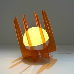 May include: A table lamp featuring a central white spherical light bulb encased within a structure of vertical, translucent orange acrylic panels. The lamp has a white power cord extending from the base and emits a warm, soft yellow glow.