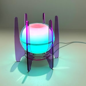 May include: A spherical lamp with a glowing pink and blue light, held within a purple acrylic frame consisting of six vertical panels arranged in a circular pattern. The lamp is connected to a thin white power cord extending to the right side.