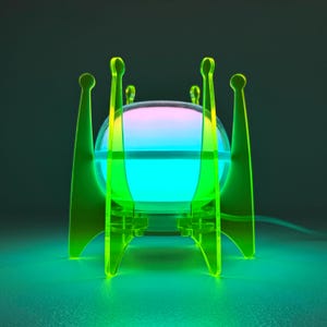 May include: A spherical lamp with a glowing gradient of pink, blue, and green light, supported by a translucent neon yellow acrylic frame featuring four tall, slender, pointed legs with rounded tips that extend upward around the central orb.