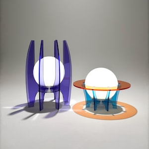 Puede incluir: Dos lámparas de mesa modernas con pantallas de globo blancas. La lámpara de la izquierda tiene una base de acrílico morado con un diseño geométrico. La lámpara de la derecha tiene una base de acrílico azul con una base naranja circular.