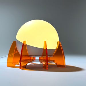 May include: A white globe lamp with a warm yellow glow sits on an orange acrylic base shaped like a rocket ship.
