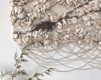 May include: A beige and gray woven wall hanging with a textured, knotted design. The hanging has a fringe at the bottom and is attached to a wooden dowel.